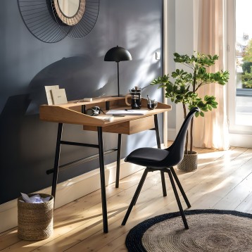 Escritorio de madera clara con patas negras y cajón integrado en ambiente de oficina moderna junto a silla negra y plantas