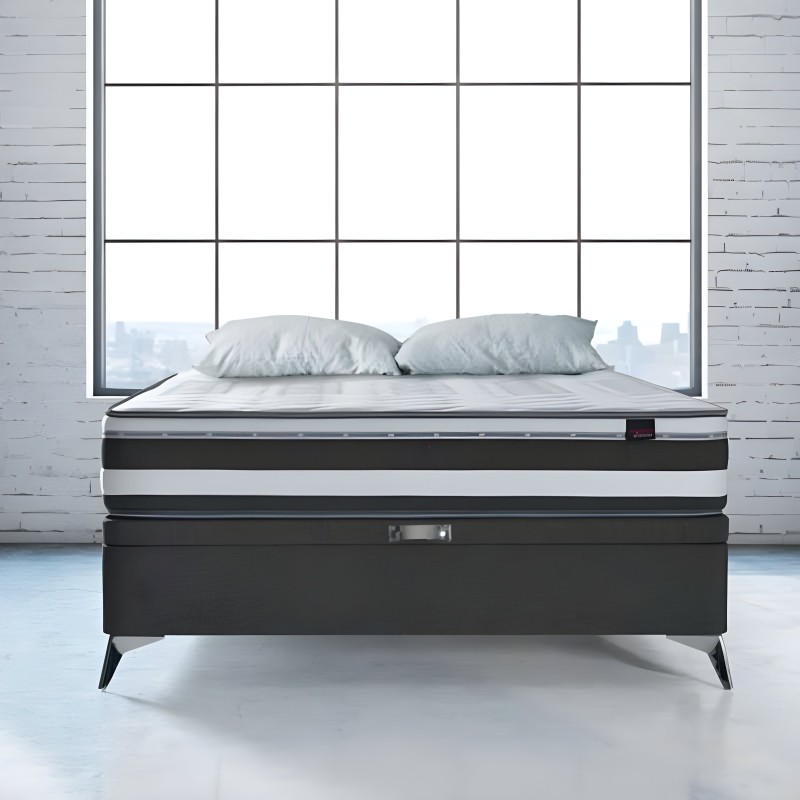 Cama con base negra y patas metálicas frente a ventanal con ladrillos blancos y dos almohadas grises