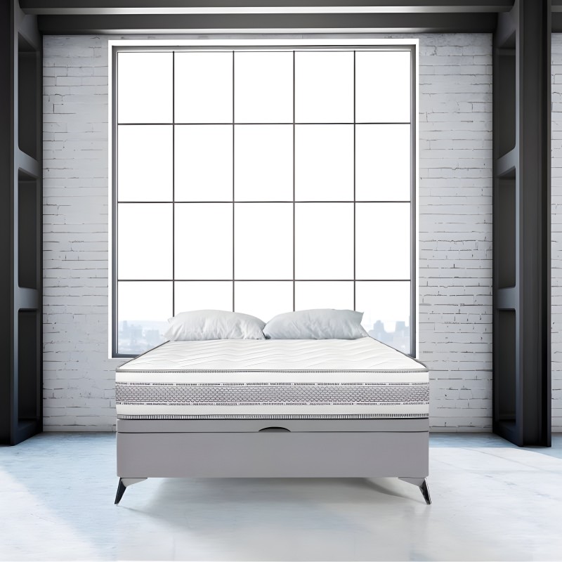 Cama tapizada gris claro con cabecero acolchado y patas metálicas en ambiente moderno junto a ventana de grid