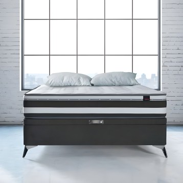 Cama con base tapizada negra y patas metálicas frente a ventanal con vistas urbanas y dos almohadas grises